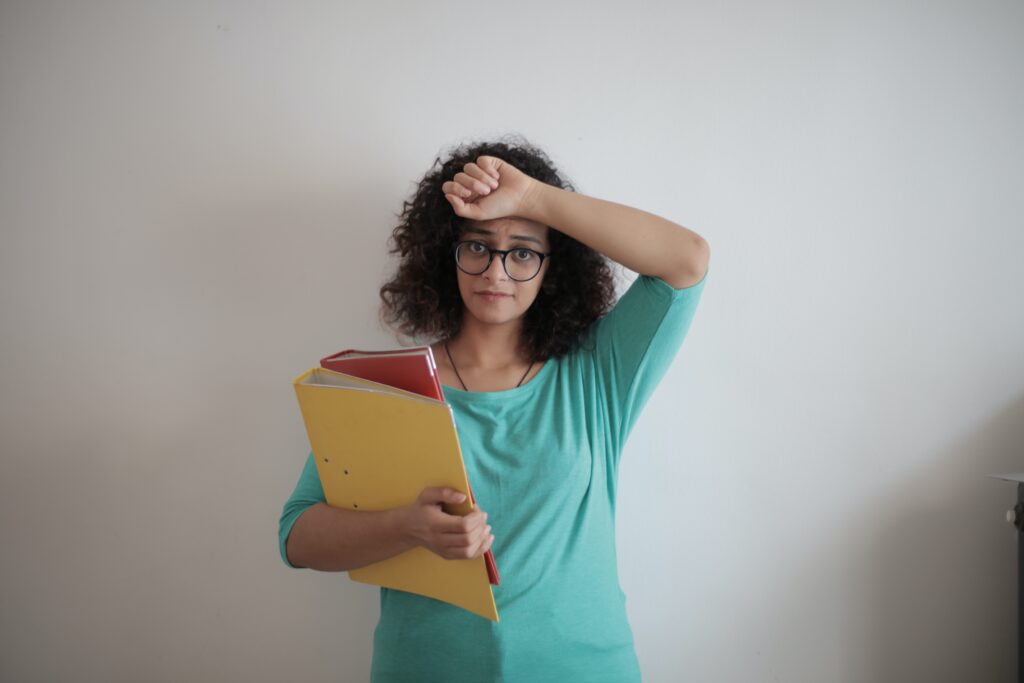 girl standing with books in her hand and arm on her forehead looking stressed and overwhelmed  